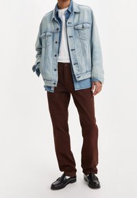Chaqueta de mezclilla azul claro con diseño clásico y botones de metal, combinada con pantalones marrones. Zapatos negros completan el conjunto.