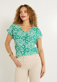 Blusa verde con estampado, escote en V y mangas cortas, con intrincados diseños en blanco. Combinada con pantalones ajustados beige.