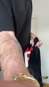 Person wearing a black shirt holds a black garment with red and white letters, extending arm toward the camera indoors.