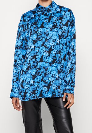 Koszula na guziki z długim rękawem w jasnoniebiesko-czarny kwiatowy wzór, noszona z czarnymi skórzanymi spodniami, ręce modela swobodnie opadające wzdłuż ciała.