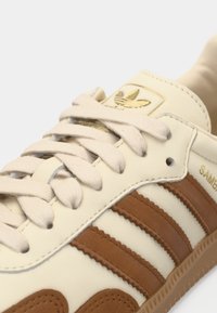 Κρεμ δερμάτινο αθλητικό παπούτσι με υφασμάτινες καφέ ρίγες, μπεζ κορδόνια και χρυσό λογότυπο στη γλώσσα. Λαστιχένια σόλα με διακριτικό μοτίβο.