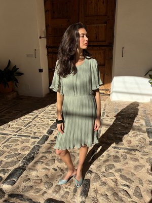 Kobieta z długimi ciemnymi włosami stoi na brukowanej nawierzchni, ubrana w jasnozieloną plisowaną sukienkę i jasnoniebieskie sandały z odkrytymi palcami, blisko drewnianych drzwi.