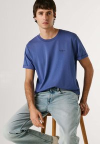 Mladý muž sediaci na drevenej stoličke, oblečený v modrom tričku s vyhrnutými rukávmi a svetlo praných džínsach na jednoliatom pozadí.