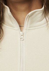 Světlý béžový zipový kabát s vysokým límcem, texturovanou látkou a výrazným detailem zipu podél přední části.