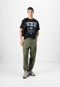 Juodas medvilninis marškinėliai su grafika "NIKE 58", alyvuogių žalios spalvos kelnes ir pilkus sportbačius; modelis laiko juodą petinę rankinę.