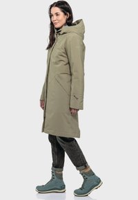 Helle olivgrüne, wasserfeste lange Jacke mit Kapuze, mit Seitentaschen und verstellbaren Manschetten, kombiniert mit leichten tealfarbenen Stiefeln.