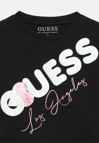 Μαύρο βαμβακερό T-shirt με μεγάλο λευκό κείμενο "GUESS", ροζ μεταλλική λεπτομέρεια και τη λέξη "Los Angeles" με καλλιγραφικά γράμματα από κάτω. Στρογγυλή λαιμόκοψη.