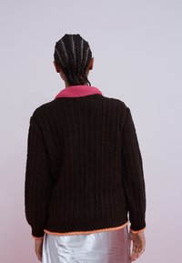 Personne aux cheveux tressés portant un pull en tricot noir avec un col rose et des bordures orange, associée à une jupe argentée brillante, vue de dos.