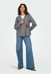 Grijze, gestructureerde wikkeljas met een striktaille, gecombineerd met wijde blauwe jeans en puntige bruine schoenen. Eenvoudig wit topje eronder.