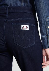Close-up of dark blue jeans pocket with white Wrangler label and visible stitched "W," worn with a medium blue denim jacket sleeve.