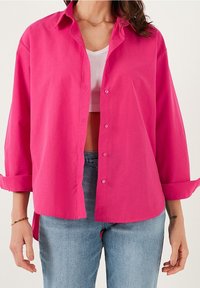Camicia rosa fucsia con colletto, maniche lunghe arrotolate sui polsini e una vestibilità ampia, indossata sopra una maglia bianca a coste.
