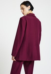 Maronroter, maßgeschneiderter Blazer mit Reverskragen, einteiligen Manschetten und einer strukturierten Passform. Glatte Textur und minimale Nähte.