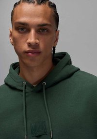 Ciemnozielona bluza z kapturem ze sznurkami, teksturowaną tkaniną i naszywką z logo. Modelka ma warkoczyki i metalową kolczyk. Zbliżenie.