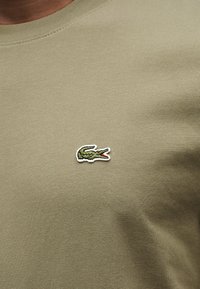 Κοντινό πλάνο σε ύφασμα χρώματος ελιάς με μικρό πράσινο κεντητό λογότυπο κροκοδείλου.