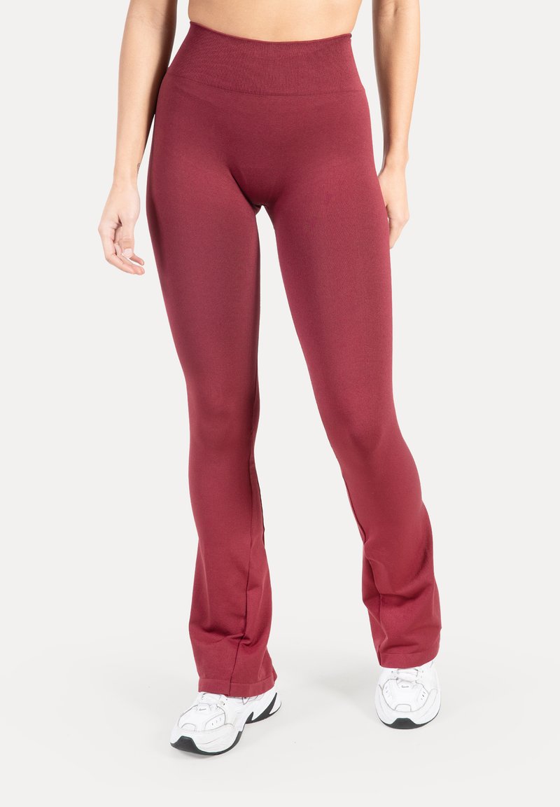 Femme portant un legging évasé taille haute couleur bordeaux et des baskets blanches de sport, debout contre un fond clair uni.