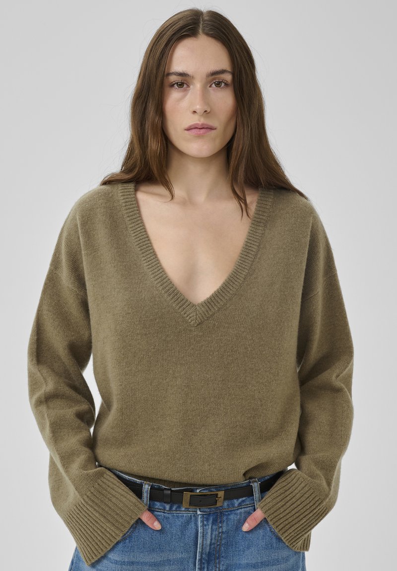 Pull col en V vert olive en tissu doux avec ourlet côtelé et manches larges, associé à un jean en denim bleu taille haute et une ceinture noire.