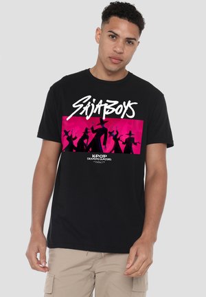 Man wearing black t-shirt with white "Sajaboys" text and pink graphic of shadowy figures in hats labeled "Kpop Demon Hunters."