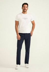 Maglietta bianca con testo nero, pantaloni blu navy e sneakers bianche. L'outfit presenta una vestibilità casual e un design minimalista.