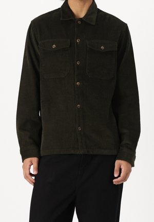 Homme portant une veste-chemise en velours côtelé vert foncé avec des boutons à l'avant et deux poches poitrine, associée à un pantalon noir.