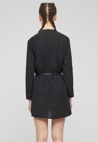 Zwarte wikkeljurk met lange mouwen, textuurstof en een riem in de taille. Het ontwerp heeft een gebogen zoom en is knielang.