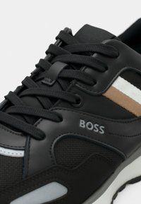 Baskets noires en matériaux mesh et cuir. Présente des accents marron et blancs, des lacets ronds, un col rembourré et une semelle en caoutchouc. Logo "BOSS" présent.