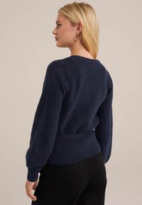 Temno modra pletena jopica z dolgimi, napihnjenimi rokavi,oprimo pasu in mehkega materiala. Zadnji pogled prikazuje gladek, zaobljen ovratnik in diskretne šive.