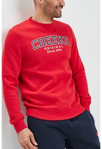 Sudadera roja con cuello redondo, que presenta "CREEKS ORIGINAL Desde 1974" impreso en azul marino y blanco. Textura suave y sólida, puños acanalados.