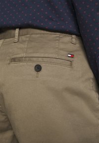 I pantaloni khaki presentano una tasca posteriore con un bottone nero e una piccola etichetta con logo nei colori blu, bianco e rosso. Tessuto dalla trama liscia, vestibilità sartoriale.
