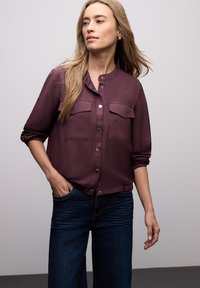 Chemise boutonnée couleur bordeaux avec deux poches poitrine, tissu léger et poignets froncés. Associée à un jean bleu foncé à jambes larges.