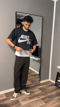 Nuori mies asettaa mustan Nike SB -paidan valkoisen paidan päälle, seisoo korkean peilin edessä modernissa huoneessa, jossa on puulattia.