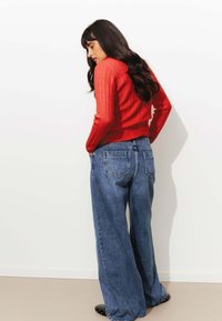 Pull en maille rouge texturé associé à un jean bleu taille haute à jambes larges avec des poches arrière. Des ballerines noires à clous complètent la tenue.