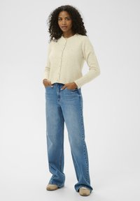 Frau mit Locken, die einen beigen Knopfcardigan, weite blaue Jeans und beigen Sneakers trägt, steht mit den Händen in den Taschen.