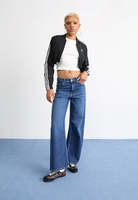 Veste noire cintrée avec des manches à rayures blanches, top ajusté blanc, jeans en denim bleu à jambe large et baskets noir-brun avec des lacets blancs.