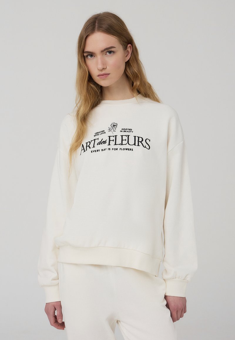Felpa di colore crema realizzata in tessuto morbido, con testo ricamato nero e motivi floreali. Maniche lunghe con polsini elasticizzati, vestibilità comoda.