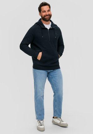 Sudadera de color azul oscuro con cuello con cremallera, bolsillo frontal y puños de canalé. Combinada con jeans de color azul claro y zapatillas grises.