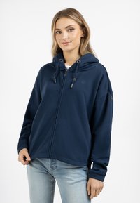 Námořnická modrá mikina na zip vyrobená z měkkého materiálu, s kapucí na šňůrku, klokaní kapsou a žebrovanými manžetami. Diskrétní logo na hrudi.