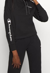Sudadera negra con un cuello alto y cordones, que presenta el logo en blanco de "Champion" en el pecho y la manga. Tejido de algodón, puños acanalados.