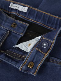 Temno modre kavbojke iz jeansa z zadrgo na sprednji strani, z zlatim šivom, z gumbnim zapiranjem in mehko notranjo podlogo, vidno na pasu. Notranja etiketa znamke.