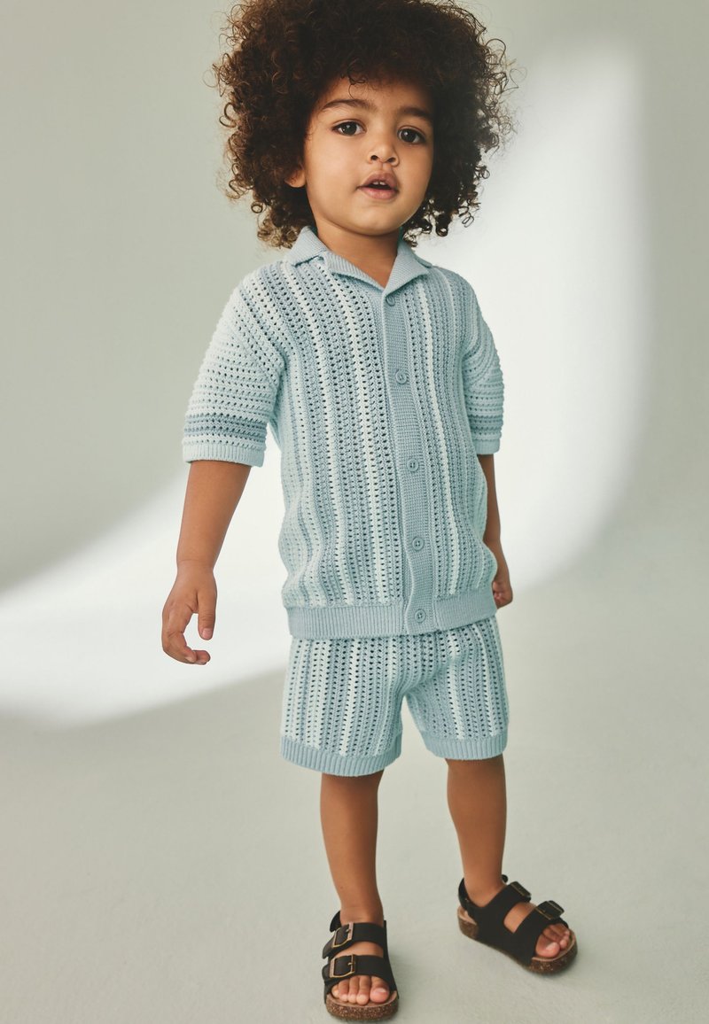 Young child with curly hair wearing light blue knit button-up shirt and matching shorts with black sandals standing on a light background.