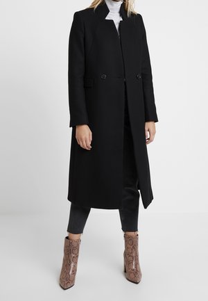 Vrouw draagt een lange zwarte jas over een witte coltrui, korte zwarte broek en beige enkellaarsjes met slangenprint, staand tegen een effen achtergrond.