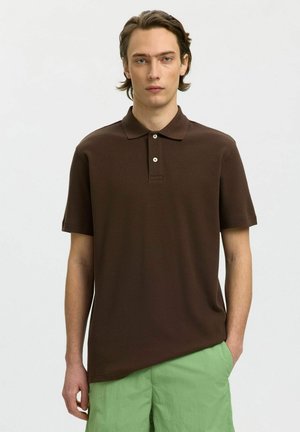 Ung mand med mellemlangt brunt hår iført en mørkebrun kortærmet polo og lysegrønne bukser, stående mod en ensfarvet baggrund.