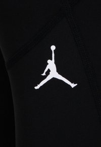 Crna tkanina glatke teksture sadrži bijeli Jumpman logo. Detaljno šivanje duž rubova dodatno ističe dizajn.