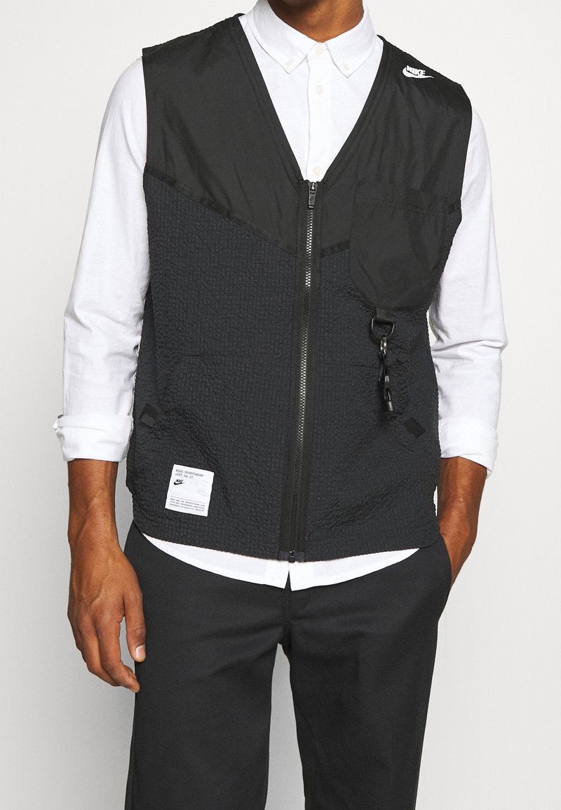 Veste noire en tissu texturé, fermeture éclair, poches latérales et un logo bien visible sur la poitrine. Portée par-dessus une chemise blanche.