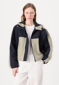Veste à capuchon bicolore avec des panneaux bleu marine et vert olive, fermeture éclair à l'avant, poignets élastiques et deux poches frontales. Tissu léger.