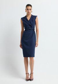 Femme debout dans une robe sans manches bleu marine ceinturée et des escarpins bruns à bout pointu sur un fond blanc uni.