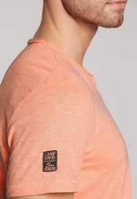 Jasnopomarańczowy t-shirt z krótkim rękawem, wykonany z miękkiego materiału, z czarną metką z białym napisem przyszytą do lewego rękawa.