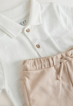 Polo bianco testurizzato con bottoni in legno abbinato a pantaloni beige con coulisse, distesi su una superficie bianca.