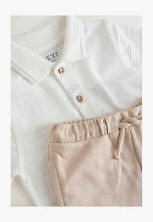 Biely textúrovaný polo tričko s drevenými gombíkmi v kombinácii s béžovými nohavicami na sťahovanie, položené na bielej ploche.