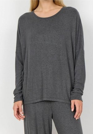Femme portant un haut ample à manches longues gris foncé et un pantalon assorti, debout devant un fond clair uni.