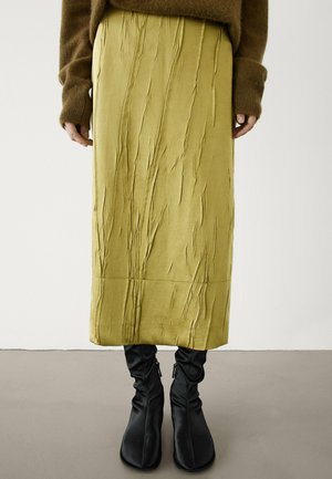 Personne portant une jupe midi vert olive froissée, un pull marron avec des poignets allongés, et des bottines noires décontractées, debout sur un sol gris.